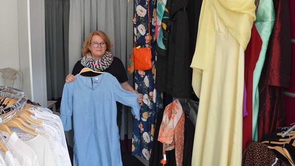 Eine Frau steht zwischen Kleiderständern und hält ein Kleid in den Händen, hinter ihr eine Umkleidekabine