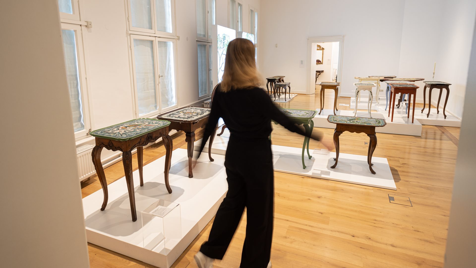 Ein Blick in die Ausstellung über die Corallenfabrik van Selow, eine Frau betritt einen Ausstellungsraum mit vielen kleinen Tischen mit Perlentischplatten