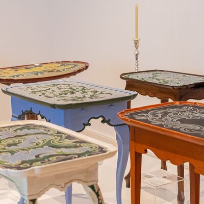 Sieben Tische in einer Ausstellung, in unterschiedlichen Farben und mit verschieden Perlenmosaiken als Tischplatte
