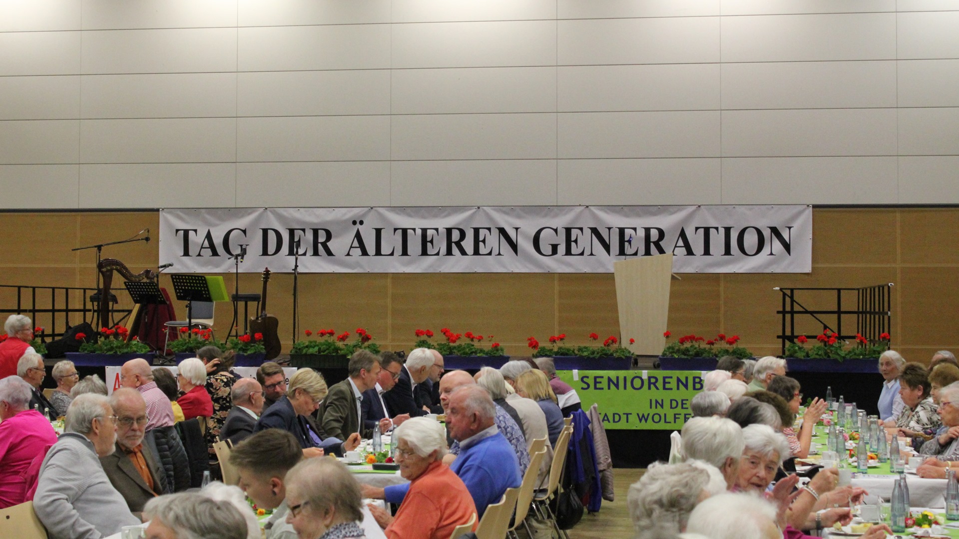 Hinter einer Bühne ist ein Banner mit der Aufschrift "Tag der älteren Generation" angebracht. Auf der Bühne steht ein Rednerpult, Mikrofonständer, eine Harfe und eine Gitarre. Vor der Bühne sitzen an langen Gruppentischen zumeist ältere Menschen, die sich unterhalten.