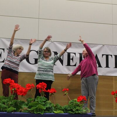 Auf einer Bühne stehen drei Frauen, die ausladende Armbewegungen machen.