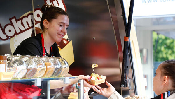 An einem Food Truck serviert eine junge Frau einer Kunding eine Schale mit Essen.