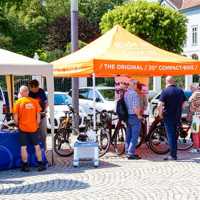 Ein Stand, Personen und Fahrräder unter einem weißen und einem orangenem Pavillon.