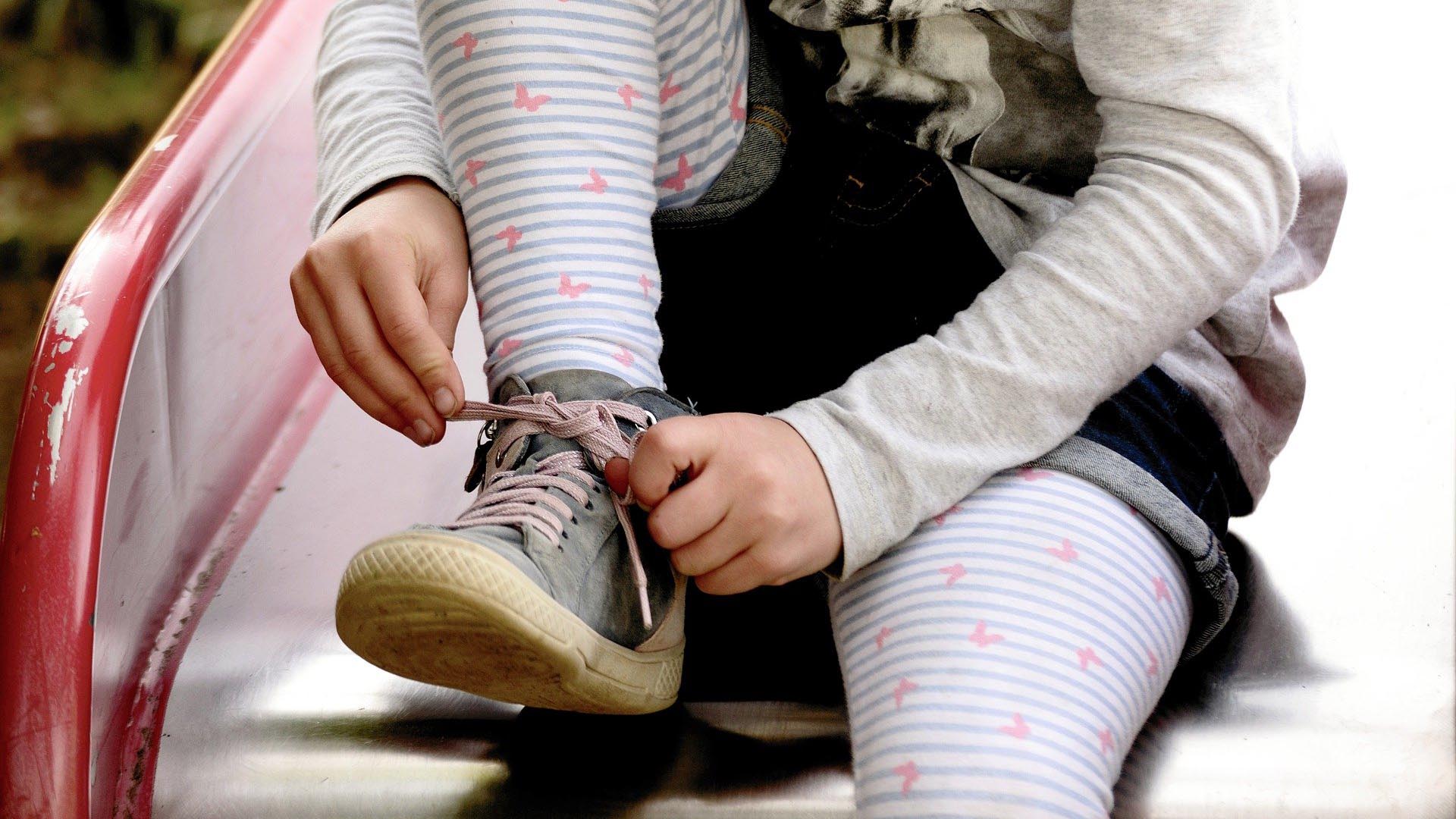 Beine und Füße eines jungen Mädchens mit rosa-blau-gemusterter Strumpfhose. Das Mädchen bindet gerade eine Schleife an ihrem Schuh.