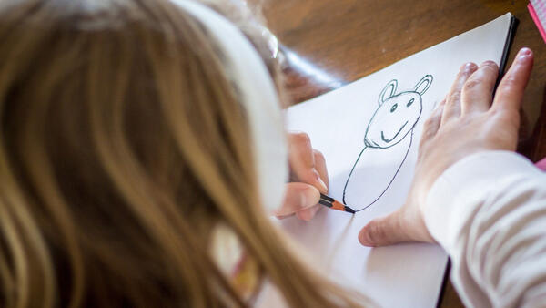 Blick über die Schultern eines kleinen blonden Mädchens, dass mit einem Stift eine Figur oder ein Tier auf ein Papier malt.