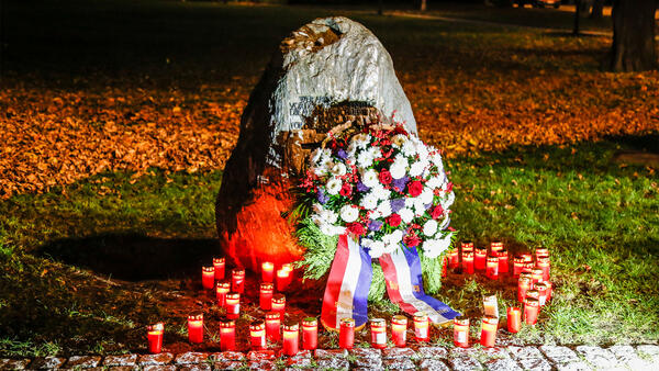 Ein einem Gedenkstein lehnt ein Kranz mit Schleife, davor stehen brennende Kerzenlichter.