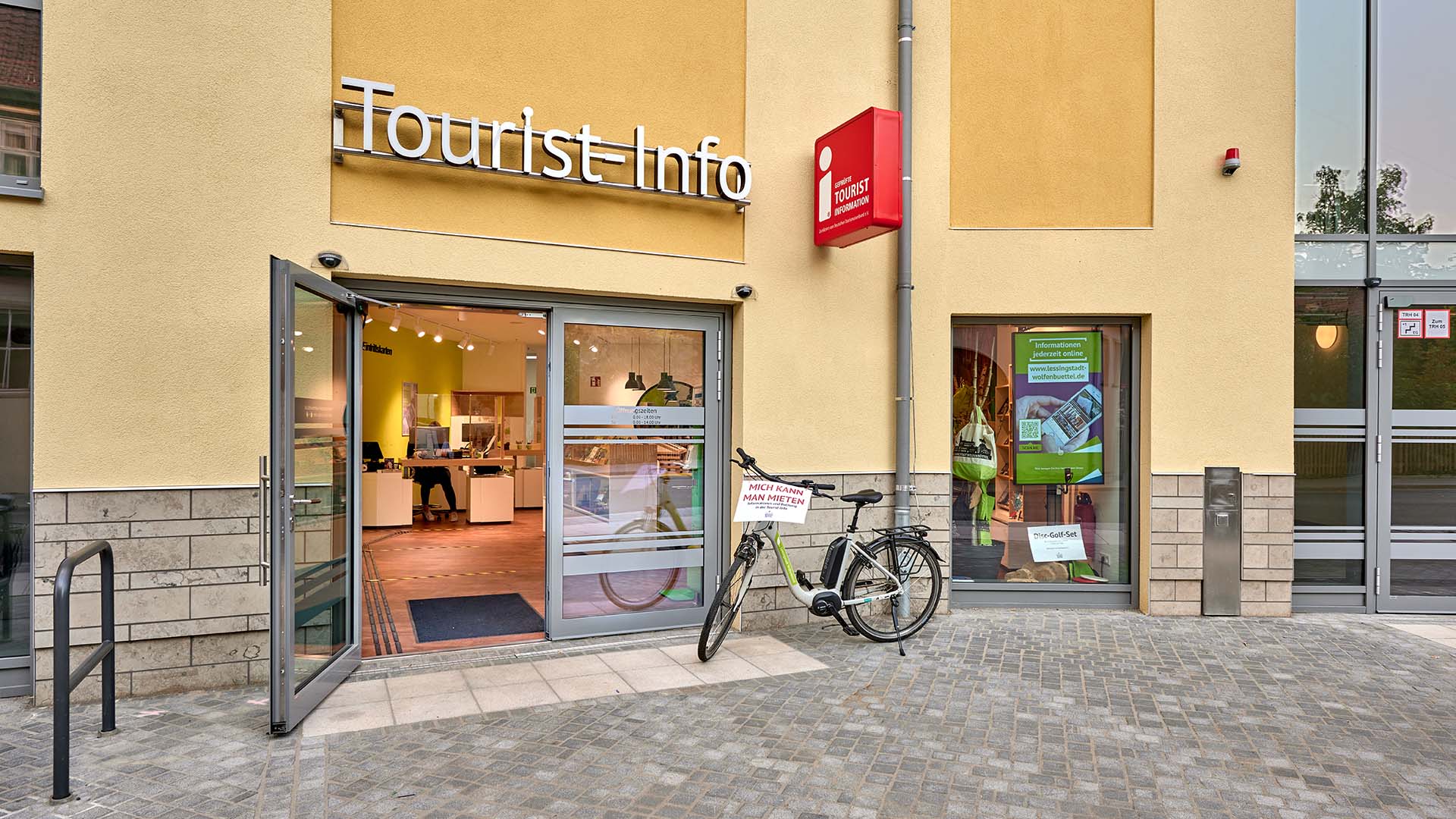 Link zur touristischen Internetseite Wolfenbüttels, Bereich Tourist-Information. - Bildmotiv: Blick auf eine Hausfront, die große Eingangstür aus Glas ist geöffnet, davor steht ein Fahrrad, das man leihen kann. Über der Tür steht Tourist-Info