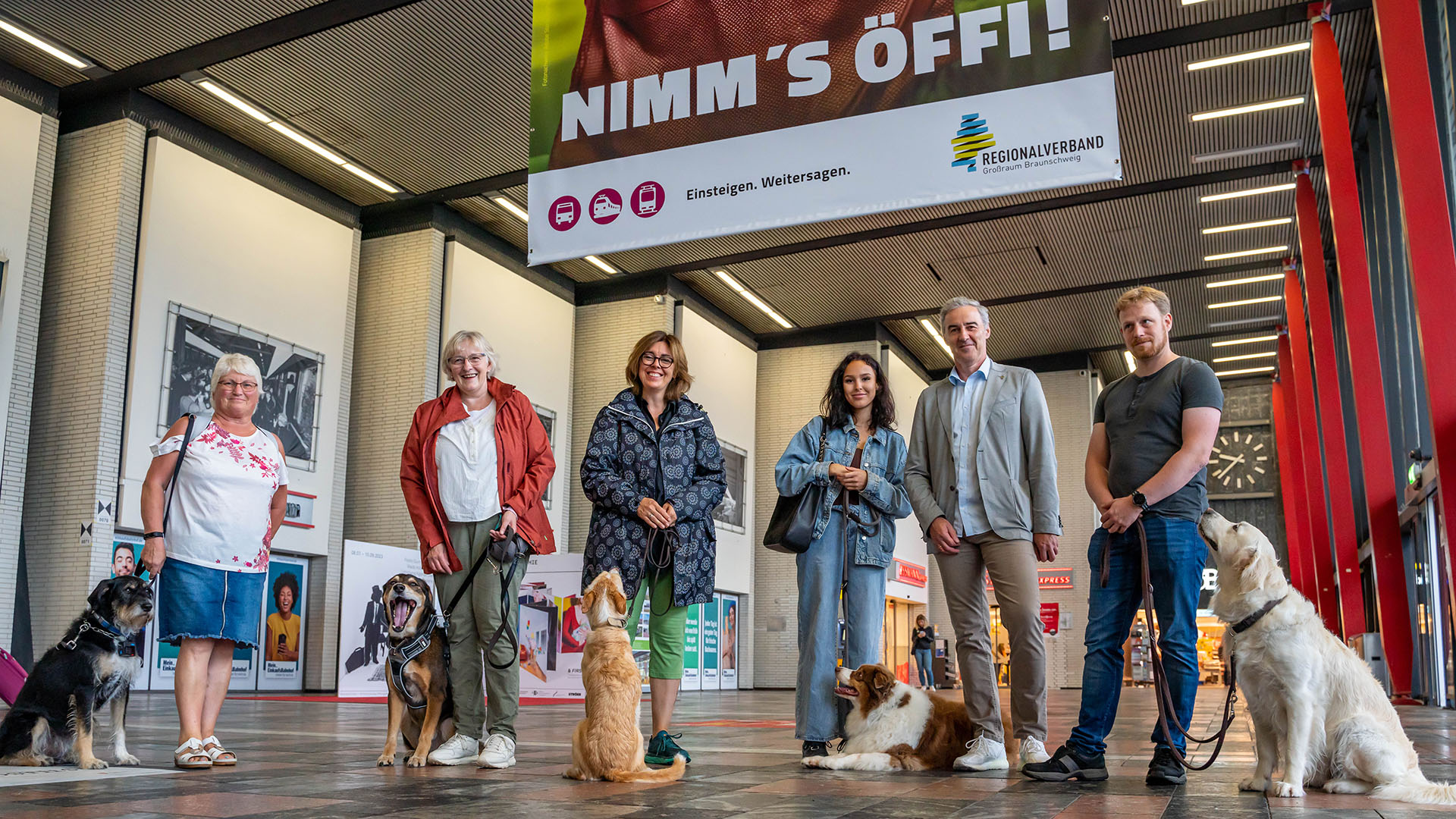 Vier Frauen und zwei Männer stehen in einer Bahnhofshalle unter einem Plakat "Nimm's Öffi!", das von der Decke hängt. Fünf Personen haben Hünde dabei.