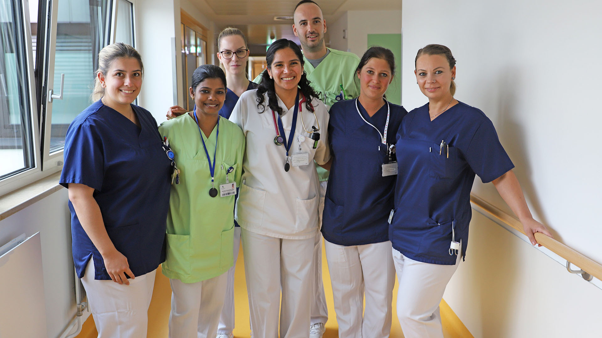 Gruppenfoto von sechs Frauen und einem Mann auf einem Krankenhausflur, alle tragen Dienstkleidung des Krankenhauses.