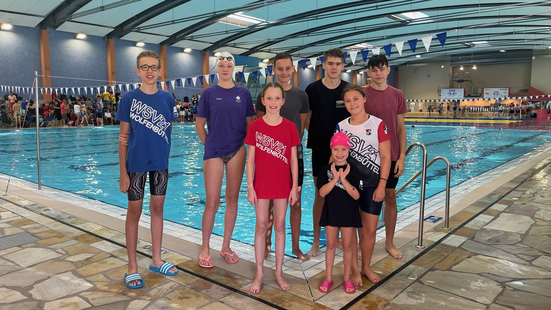 Acht Jungen und Mädchen verschiedenen Alters stehen vor einem Schwimmbecken in einem Hallenbad