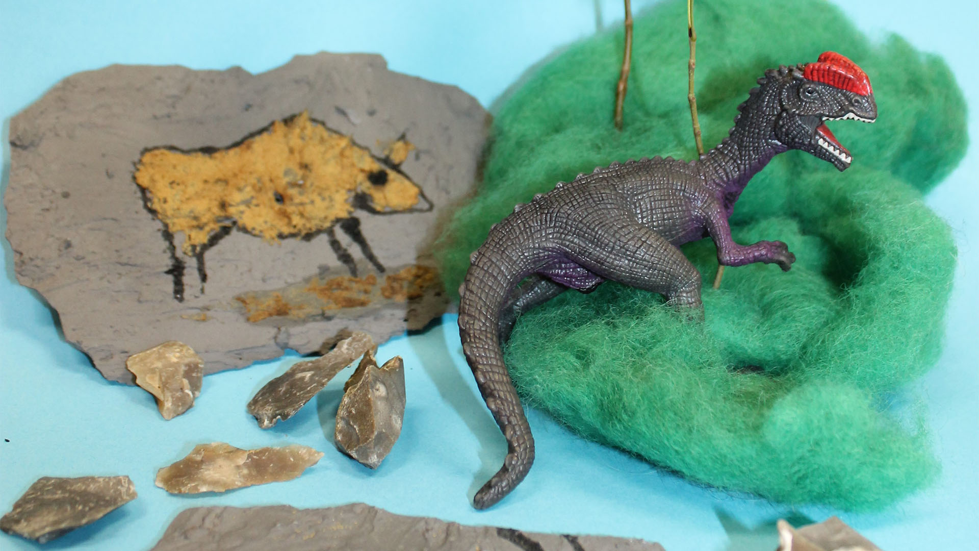 Ein Stein ist mit einem Tier bemalt daneben liegen ein paar Feuersteine. Auf etwas Wolle liegt ein grauer Spielzeugdinosaurier.