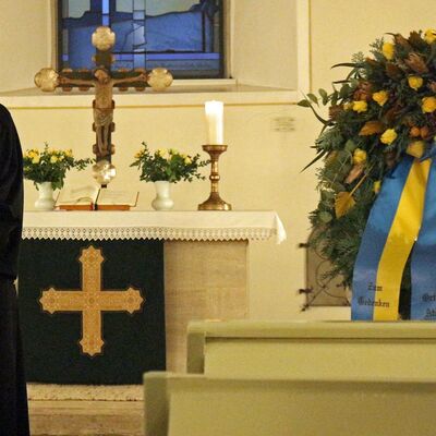 Eine junge Frau hält eine Predigt vor einem Altar mit Kreuzen, Kerzen und Blumen. Neben ihr steht ein geschmückter Kranz aus Tannenzweigen. An diesem hängen zwei blau-gelbe Bänder mit der Aufschrift "Zum Gedenken" und "Ortsrat Atzum".