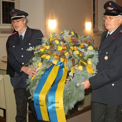 Zwei Männer in Feuerwehruniform tragen einen geschmückten Kranz aus Tannenzweigen. An diesem hängen zwei blau-gelbe Bänder mit der Aufschrift "Zum Gedenken" und "Ortsrat Atzum".