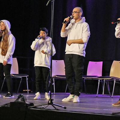 Vier junge Menschen stehen, einheitlich schwarzweiß gekleidet, auf einer Bühne und halten Mikrofone vor den Mund