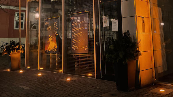Die Glasfassade des Rathausanbaus wird abends bei Dunkelheit durch Bodenstrahler orange angestrahlt. In den Fenstern hängen Veranstaltungsplakate des Theaters.