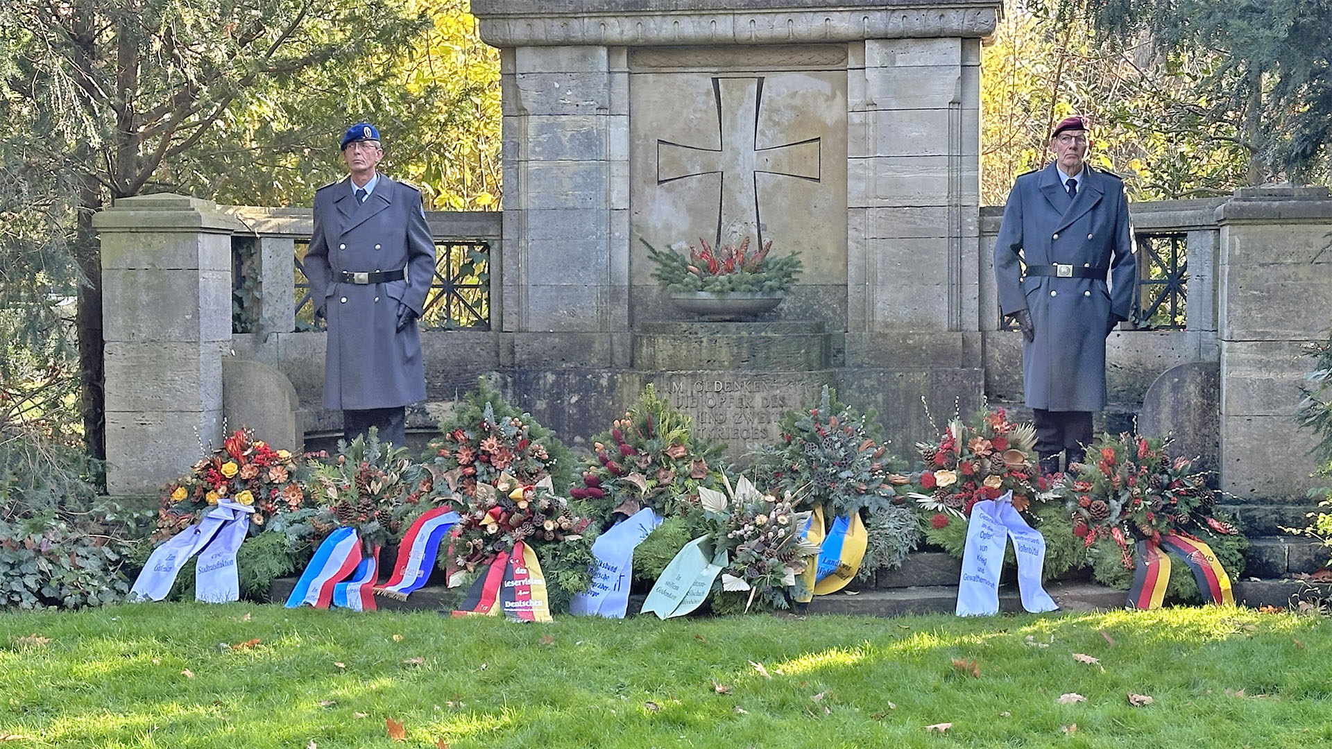 An einem Gedenk- oder Ehrenmal wurden Kränze neidergelegt, rechts und links neben dem Stein stehen jeweils ein Soldat in Uniform