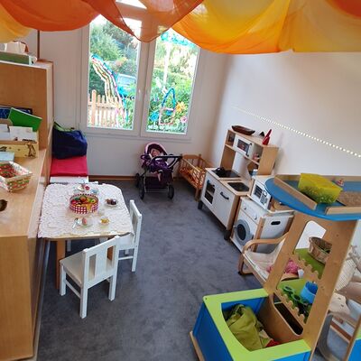 Ein Kitaraum mit einer Kinderküche, -waschmaschine und weiterem Holzspielzeug und einem Kindertisch mit -stühlen.