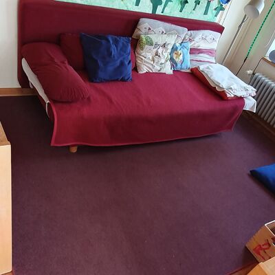 In einem Kita-Raum steht ein Sofa mit Decken und Kissen, gegenüber ein Schrank mit Spielzeutg und einer mit Obst und Getränkekannen