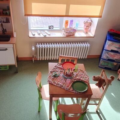 Ein Raum in der Kita mit einer Kinderküche, einem mit Tellern gedeckten Kindertisch und einem Puppenbett sowie Spielzeug