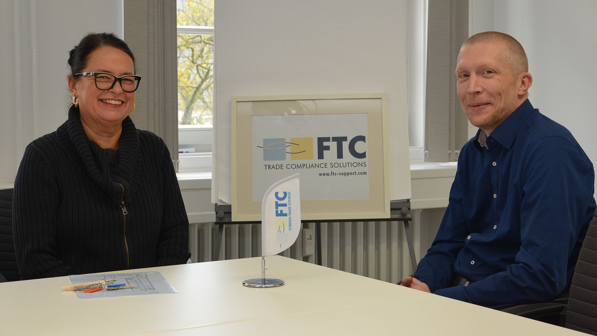 Eine Frau und ein Mann sitzen zusamnmen an einem Tisch, auf dem Tisch und im Hintergrund das Logo FTC Trade Compliance Solutions