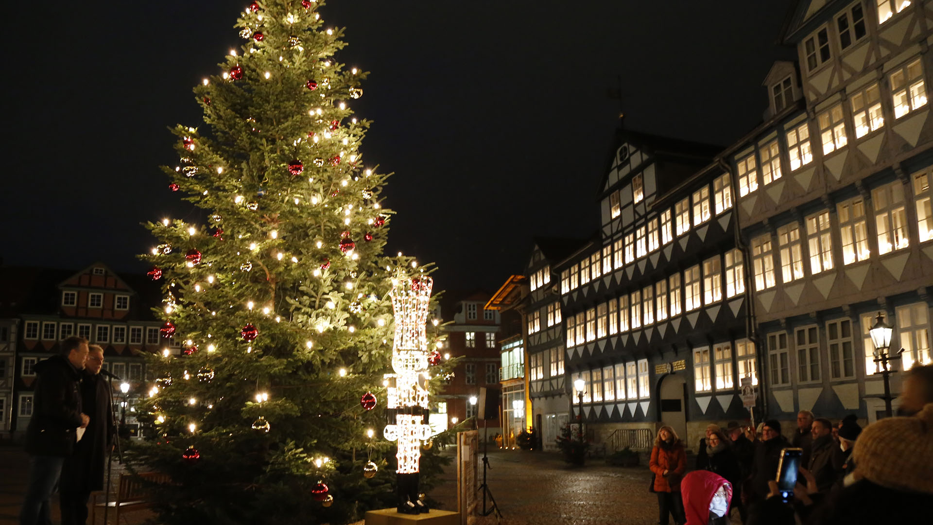 Eine große Tanne mit vielen leuchteten Lichtern steht vor dem Wolfenbütteler Rathaus. In jedem Fenster stehen leuchtende Lichterbögen. Neben der Tanne stehen zwei Männer, davor einige Menschen.