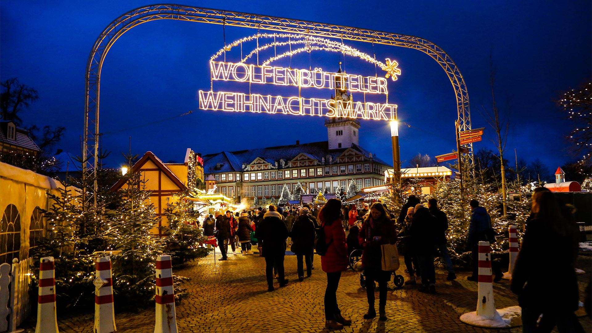 Der Eingang zum Weihnachtsmarkt, über dem der Schriftzug Wolfenbütteler Weihnachtsmarkt hängt. Man sieht viele beleuchtete Tannen, Verkaufsbuden, Menschen, im Hintergrund das Wolfenbütteler Schloss