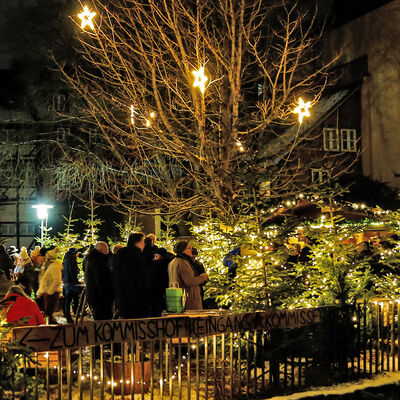 Ein Platz zwischen vielen Gebäuden mit einem beleuchteten Baum und einem beleuchteten Stand. Auf dem Hof stehen viele Personen an Stehtischen.