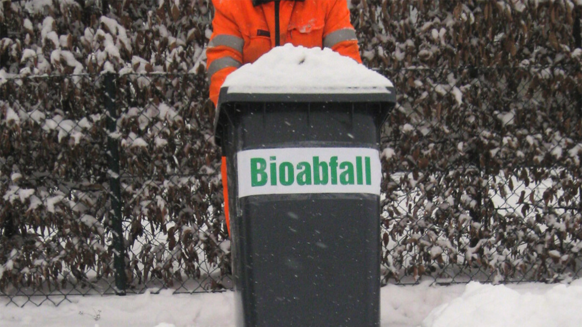 Ein Mann in oranger Arbeitskleidung zieht eine schneebedeckte Bioabfalltonne durch Schnee
