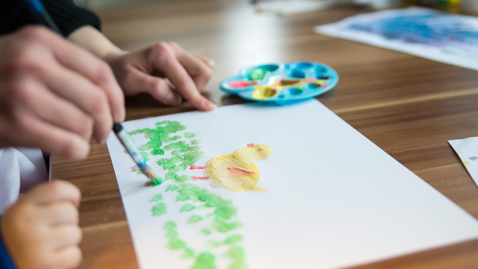 Kinderhand und die Hand einer erwachsenen Frau malne an einem Bild mit Rasen und Kücken.