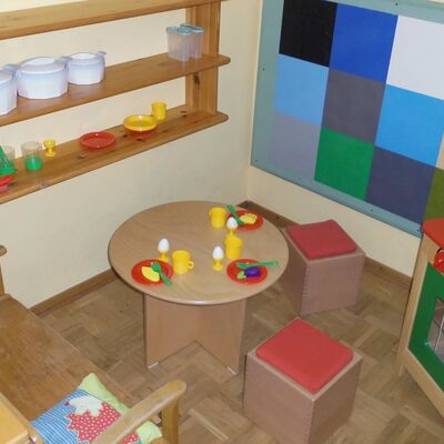 Eine kleine Kinder-Kchenecke, mit einem Herd aus Holz, einem Wandregal mit Kunststoff-Tpfen und -Geschirr, einem kleinem Tisch mit Kunststoff-Gedecken und  Sitzgelegenheiten