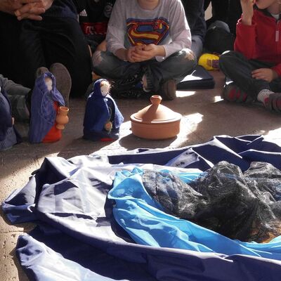 Kinder sitzen in einem Kreis, vor ihnen liegen Stoffe, die eine Landschaft nachbilden. Drei kleine Puppen und eine Puppen-Htte stellen eine Szene nach.