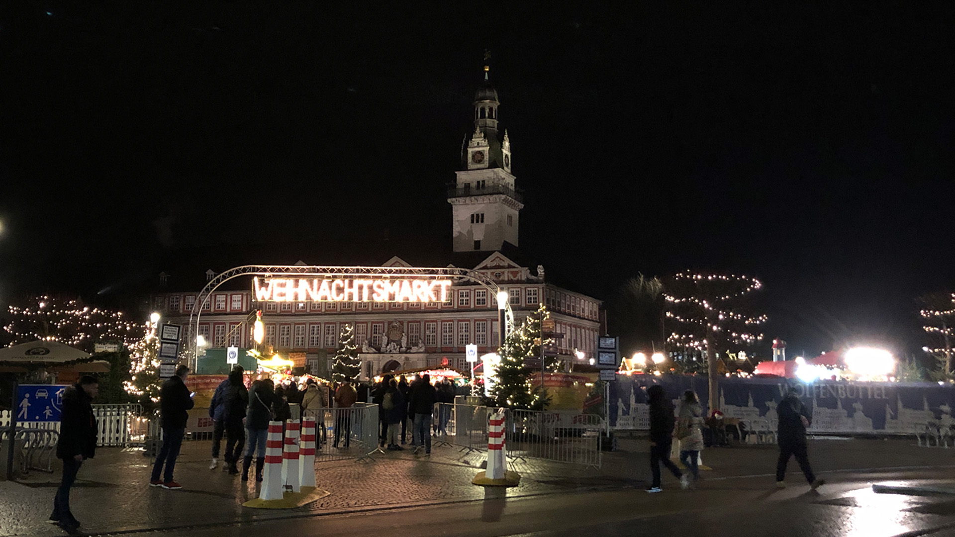 Im Dunklen leuchtet der Schriftzug Weihnachtsmarkt über einigen Verkaufsständen auf der Weihnachtsmarkt, im Hintergrund erkennt man das Wolfenbütteler Schloss. Der Weihnachtsmarkt ist eingezäunt, der Eingang mit rotweißen Pfeilern markiert.