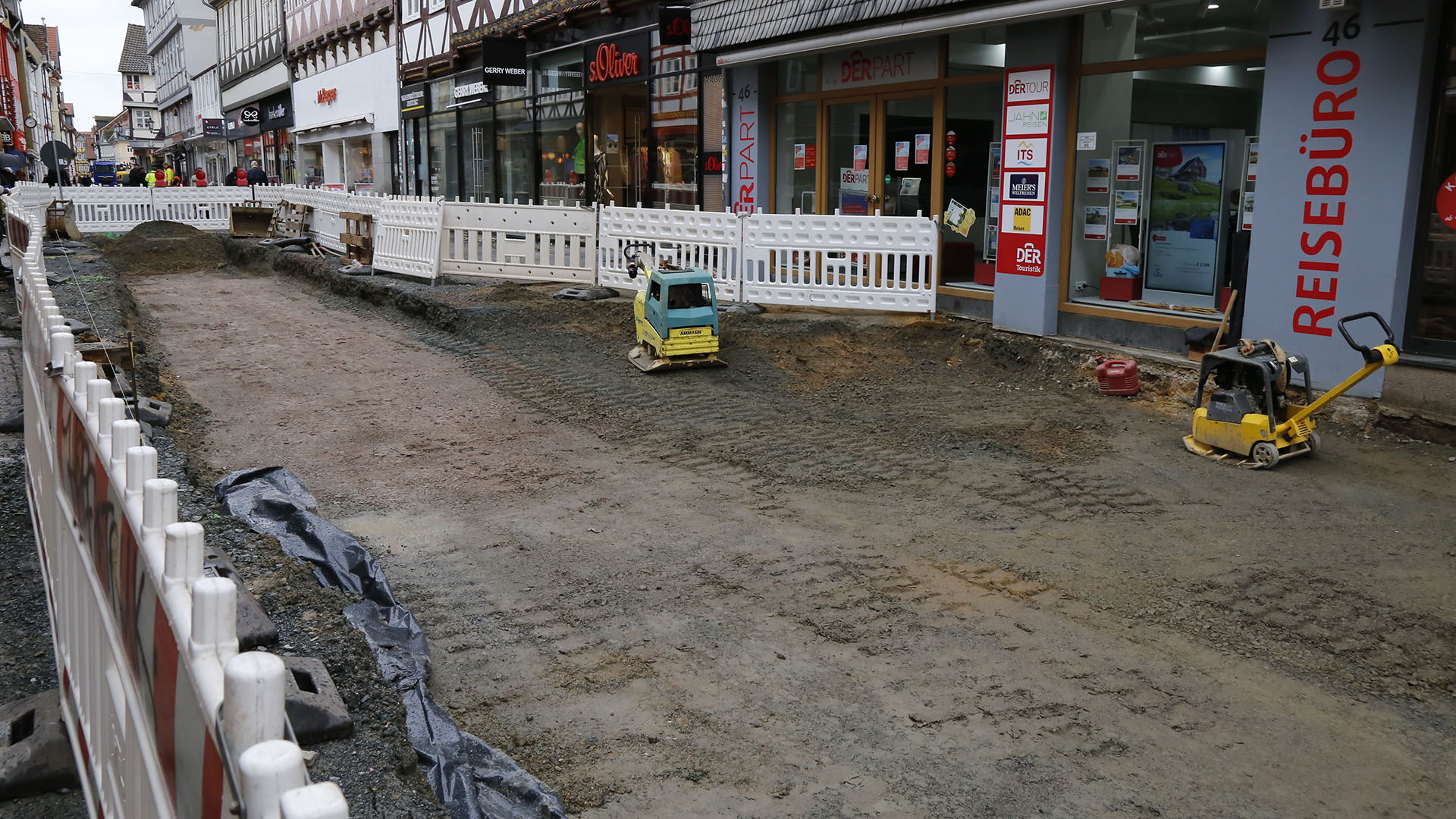 Bild vergrößern: Der mittlere Teil der Fußgängerzone zwischen den Geschäften ist mit Baustellenbarken abgesperrt, rechts und links daneben ist ein Weg für Fußgänger. In der Absperrung sieht man einen zugeschütteten Baugraben