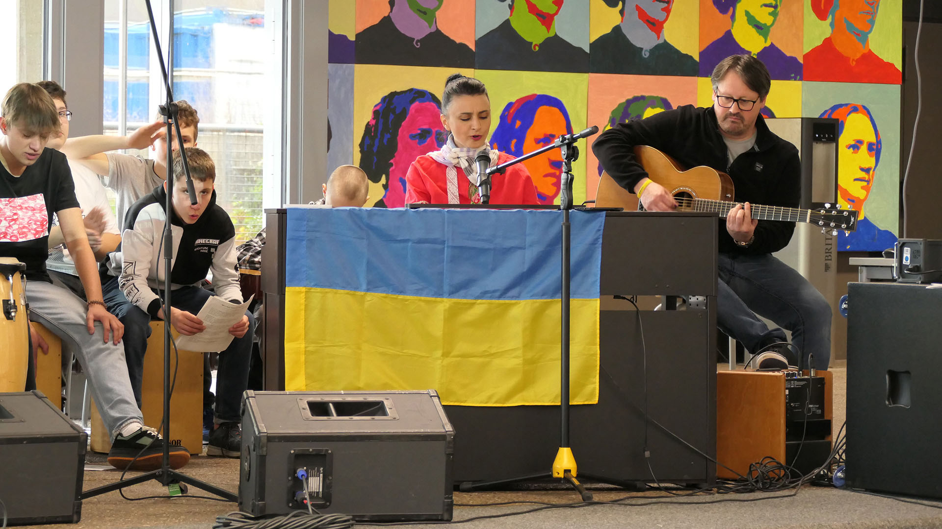 Ein Chor von Schülern sthet auf einer Tribüne und singt, im Hintergrund wird ein Text auf eine Leinwand gebeamt.