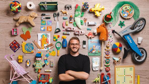 Ein junger Mann steht vor einer Wand, an der sehr viel verschiedenes Kinderspielzeug angebracht ist.