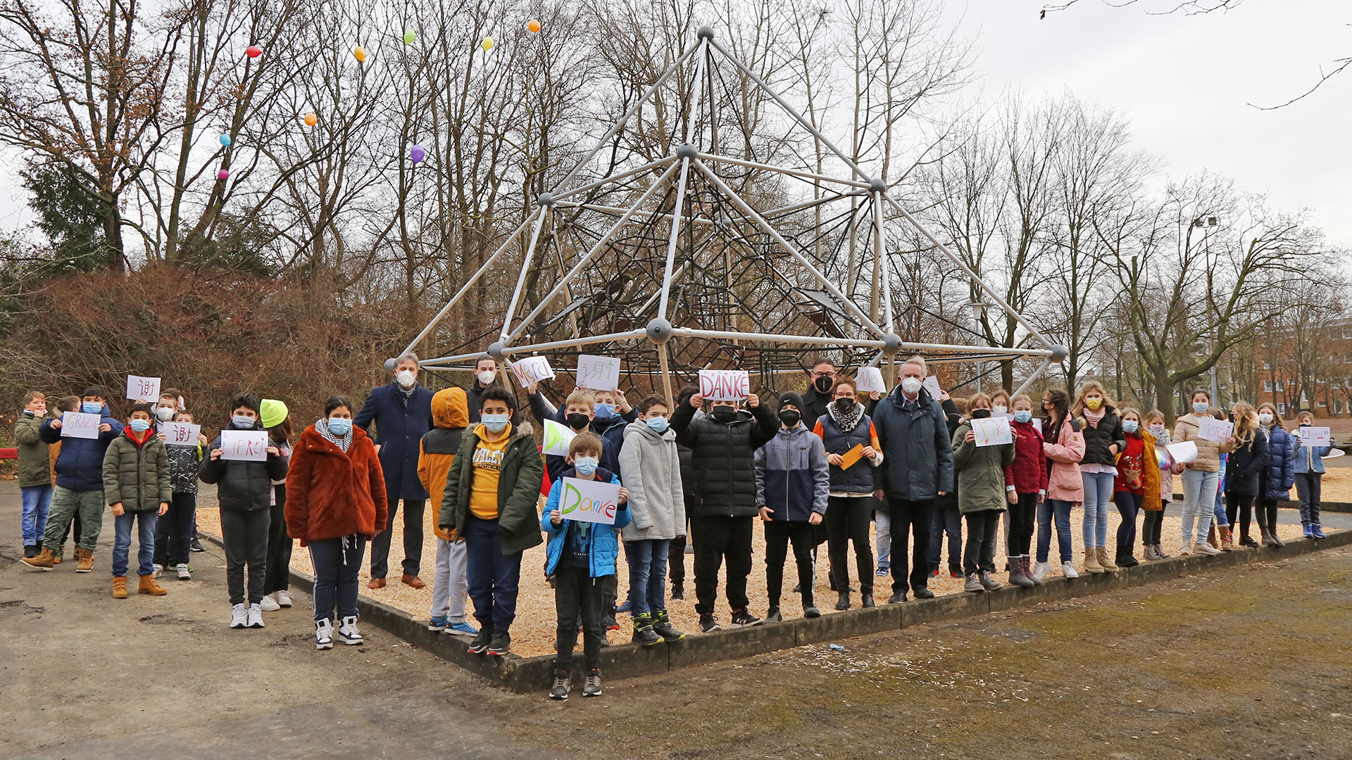 Auf einer Sandfläche eines Schulhofes steht ein großes Klettergerüst aus Metall. Davon haben sich Schülerinnen und Schüler aufgestellt, die meisten tragen Schilder "Danke" in den Händen.
