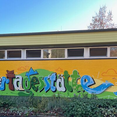 Auf einer gelb-grünen Fassade wurde mit bunter Farbe das Wort Kindertagesstätte gemalt.