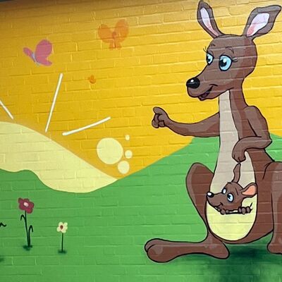 Auf einer gelb-grünen Fassade wurde im Comic-Stil ein Känguruh mit einem kleinen Känguruh im Beutel gemalt. Daneben wurden ein paar Blumen und Schmetterlinge gemalt.