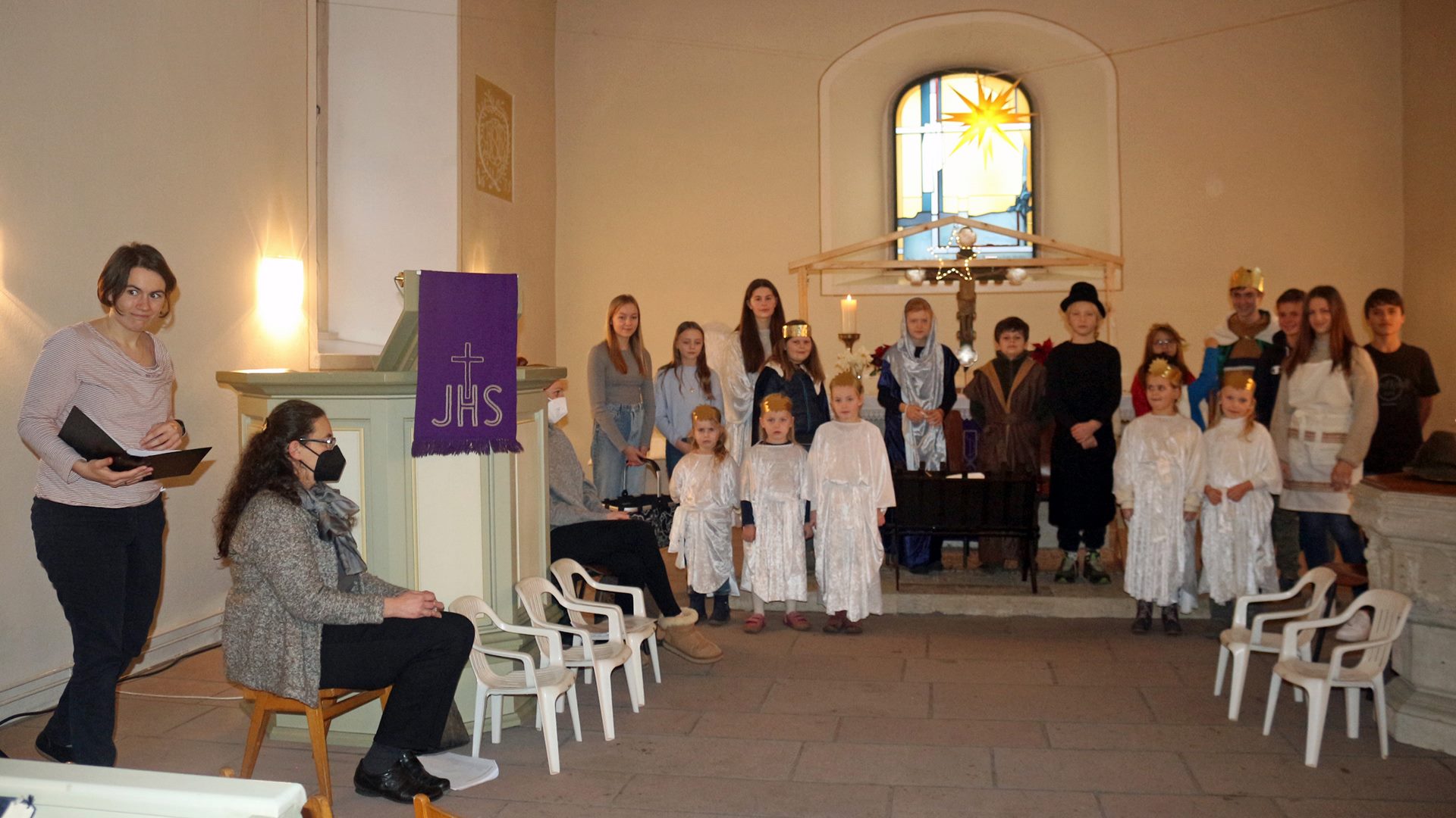 Gruppenbild von verkleideten Kindern in einer Kirche. Links im Bild befinden sich zwei Frauen. Rechts und links stehen kleine weiße Stühle.