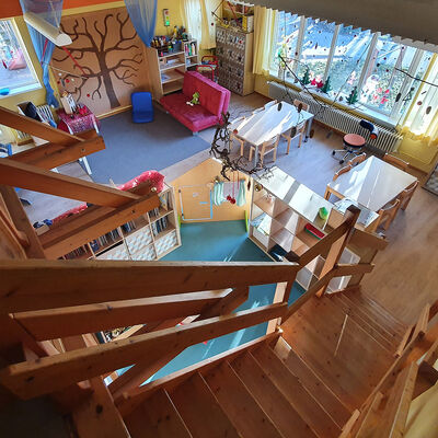 Blick von oben auf eine Holztreppe mit Geländer, die in einen Raum runterführt, in dem Tische, Sitzgelegenheiten, Regale und Spielzeug zu erkennen ist.