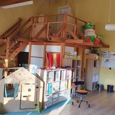 Blick auf eine Holztreppe, die in einer höhergelegene Etage auf ein Podest führt. Unter dem Podest und neben der Treppe stehen Regale mit Spielzeug.