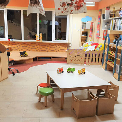 In einem Zimmer steht ein Tisch mit Stühlen in kindgerechter Grüße, an den Wänden sind Regale mit Spielzeug, in einer Zimmerecke ein Podest aus Holz.