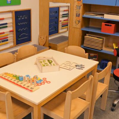 In der Technik: Ein Holztisch mit Holzstühlen und etwas Spielzeug Links an der Wand hängt ein bunter Abakus und eine kleine Tafel. Rechts steht ein Regal mit Kisten.