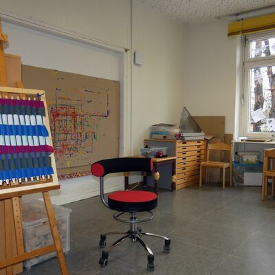 Im Atelier: Ein Raum mit großen Fenstern und grauem Fußboden. An der Seite steht ein Holztisch mit Holzstühlen. Im Vordergrund steht eine Staffelei mit etwas buntem gebasteltem. Davor steht ein rot-schwarzer Stuhl auf Rollen. An der linken Wand hängt ein großes bemaltes Banner.