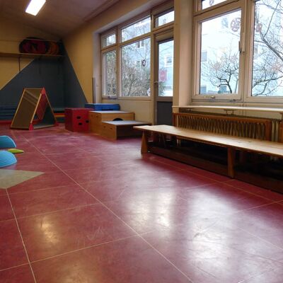 Im Bewegungsraum: Ein Raum mit großen Fenstern und rotem Fußboden. Es sind verschiedene Turn- und Spielgeräte und Bänke zu sehen.