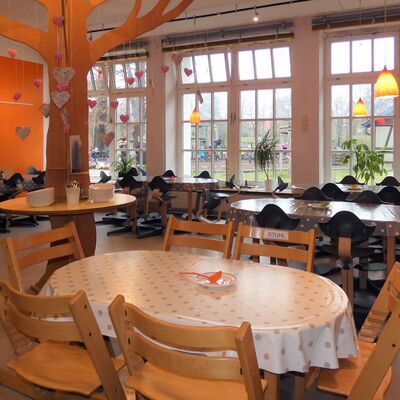 In der Cafeteria: Ein Raum mit großen Fenstern. Ein paar Tische mit Holzstühlen. Ein Stehtisch auf dem ein großer Baum aus hellem Holz aufgesetzt wurde. An dem Baum hängt an Fäden verschiedene Deko in Herzform. Eine Wand ist orange bemalt.
