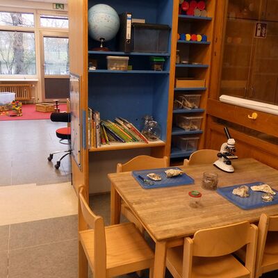 In der Technik: Rechts steht ein Holztisch mit fünf Holzstühlen. Auf dem Tisch steht ein Mikroskop. Auf Tischsets liegen Steine.
Dahinter steht ein Holzregal mit blauer Innenseite. Im Regal stehen eine Weltkugel und mehrere Bücher. Im Hintergrund ist der Rest des Raumes mit Stühlen und Spielzeug zu sehen.